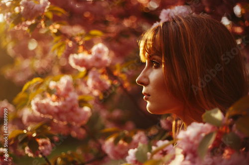 a red-haired girl with fringe looks curiously while standing between the flowers of a blossoming tree