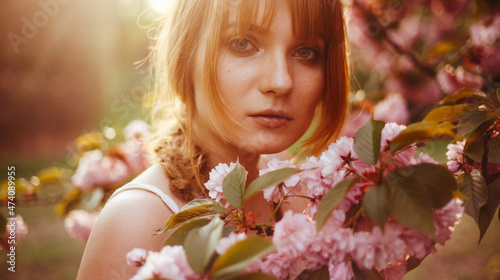 a red-haired girl with fringe looks curiously while standing between the flowers of a blossoming tree