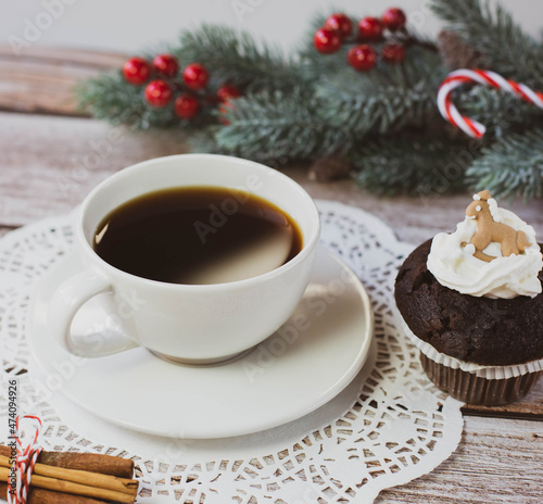 A white cup with black coffee, chocolate muffin and other Christmas decorations are on a light wooden table. Square image.	
