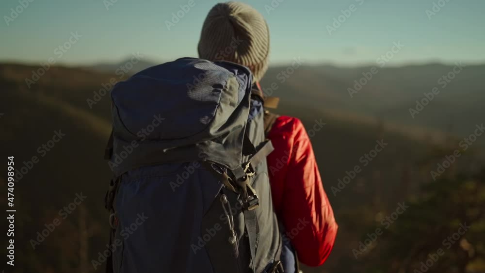 slow motion view from behind adventurous female hiker backpacker in red jacket with backpack stands on mountain top at sunset above the woods. slow motion view from behind adventurous female hiker backpacker in red jacket with backpack stands on mountain top at sunset above the woods.