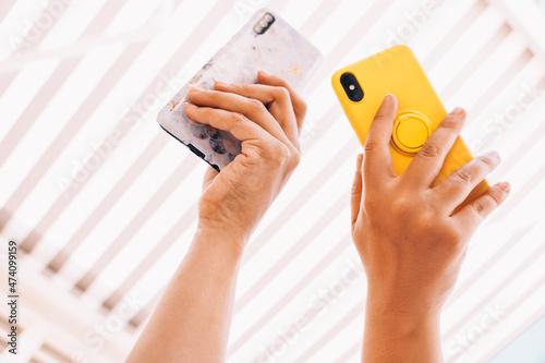 Two women using mobile phones during a sunny day, under an awning. Colorful mobiles together, sharing information. Close up focus of hands and gadgets view from below, without faces. 