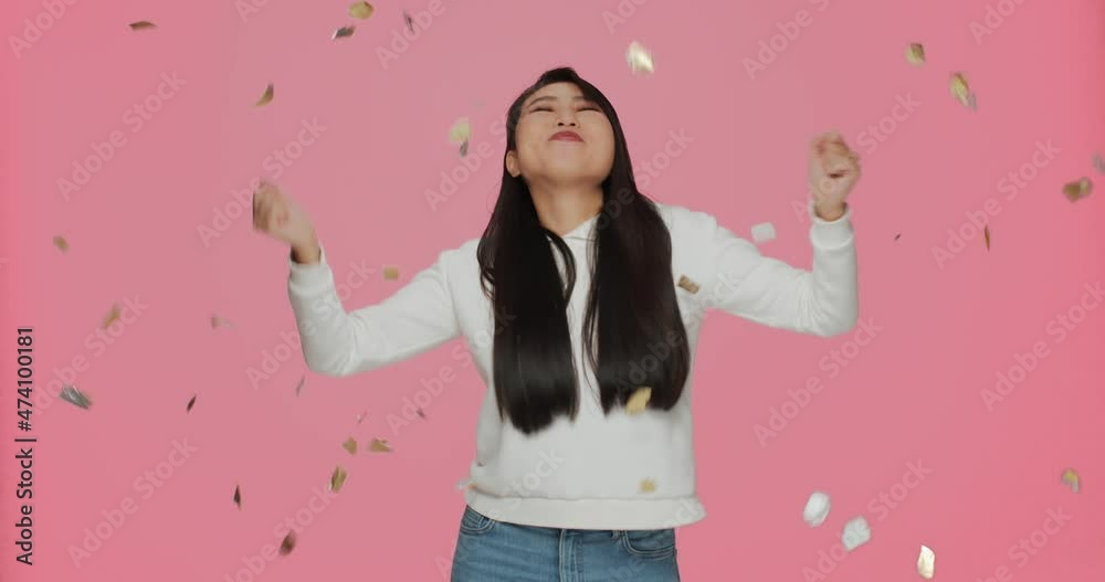 Smiling asian young girl having fun under golden confetti, celebrating success, holiday on pink studio background Smiling asian young girl having fun under golden confetti, celebrating success, holiday on pink studio background