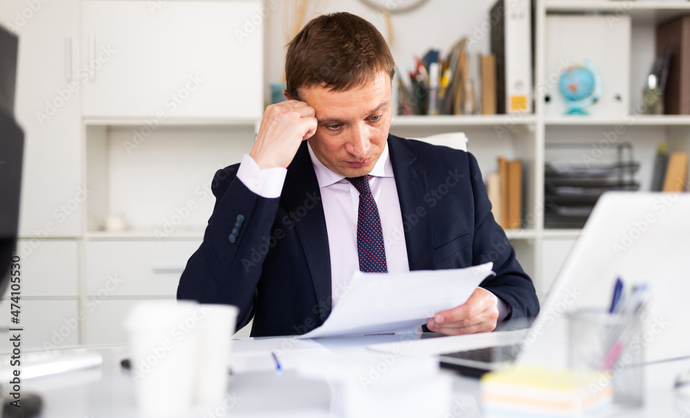 Pensive adult businessman sitting at office desk, working with papers..