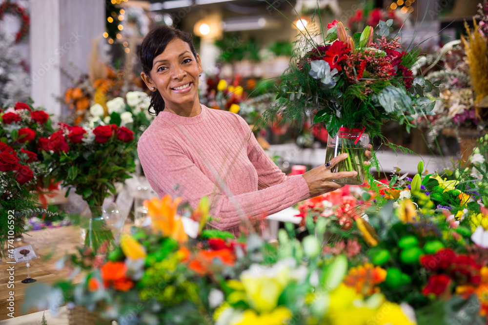 Hispanic woman standing in showroom of floral shop with bouquet of fowers in hands and looking in camera.