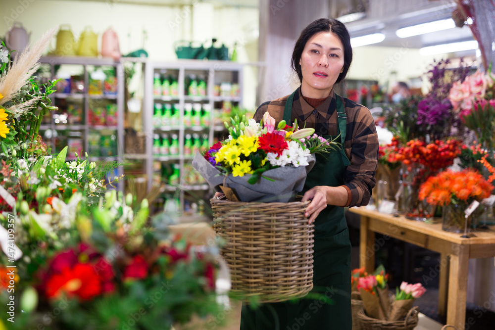 Asian woman florist carrying basket with flowers in salesroom of floral shop.