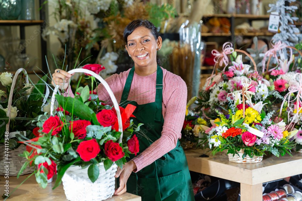 Latin woman florist standing in salesroom of floral shop and holding basket with flowers.