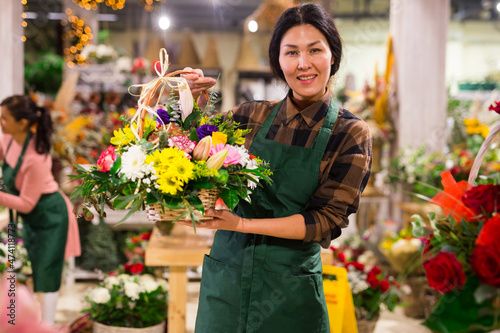 Asian woman in apron standing in showroom of floral shop and holding bouquet of flowers in hands. Her coworker Latin woman doing her job in background.