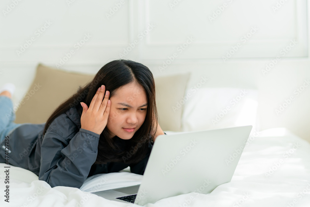 Portrait, a cute Asian teenage girl. wearing gray shirt lying in bed with a notebook computer in the bedroom to study online with a thoughtful face Portrait, a cute Asian teenage girl. wearing gray shirt lying in bed with a notebook computer in the bedroom to study online with a thoughtful face
