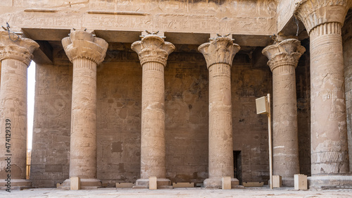 Colonnade in the ancient Egyptian temple of Horus in Edfu. Tall columns and walls are covered with carved drawings. Elegant capitals of different shapes are visible.