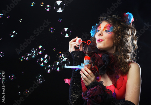 Fashionable young model female with creative make-up blowing soap bubbles. Doll style. Bright dress. Black background.