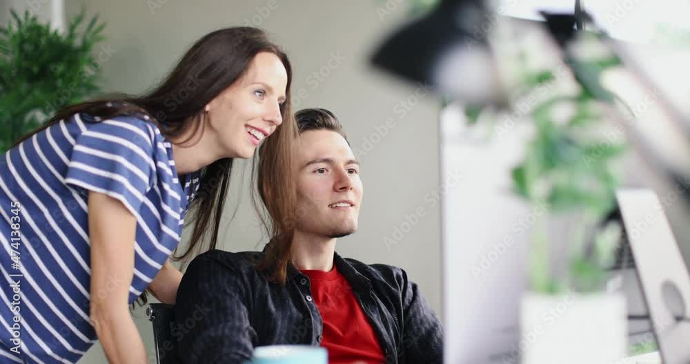 Millennial Caucasian young professional successful female worker in casual outfit standing helping discussing teaching talking with male officer colleague sitting working with computer in company. Millennial Caucasian young professional successful female worker in casual outfit standing helping discussing teaching talking with male officer colleague sitting working with computer in company.