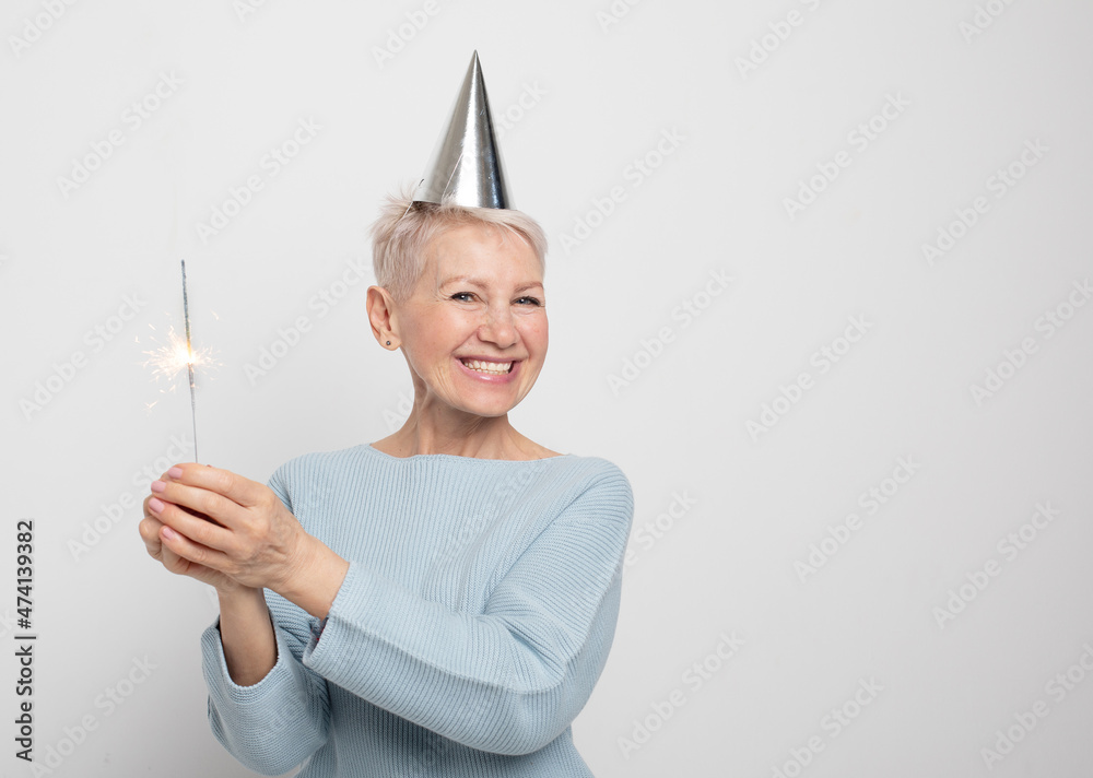 Portrait of a senior woman in studio on a gray background. Woman wearing blue sweater and party hat, holding sparkles. Portrait of a senior woman in studio on a gray background. Woman wearing blue sweater and party hat, holding sparkles.