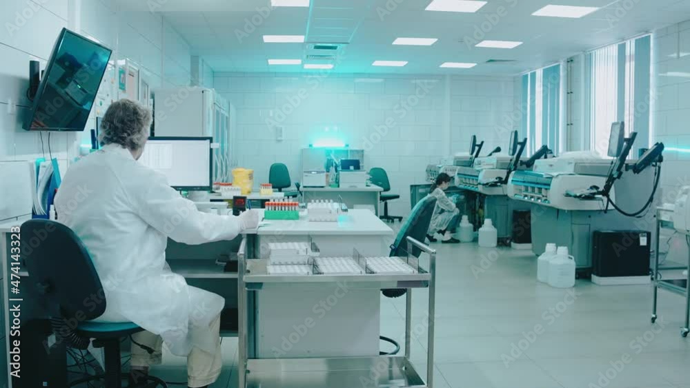 Elderly doctor sits in a laboratory with test tubes and works on a computer. Male laboratory assistant with gray hair working with documents. Work in the laboratory. High quality 4k footage Elderly doctor sits in a laboratory with test tubes and works on a computer. Male laboratory assistant with gray hair working with documents. Work in the laboratory. High quality 4k footage
