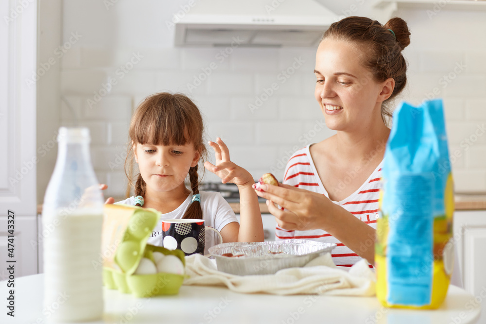 Portrait of optimistic happy woman sitting at table with her daughter and eating tasty cakes, posing in kitchen, expressing positive emotions and happiness.