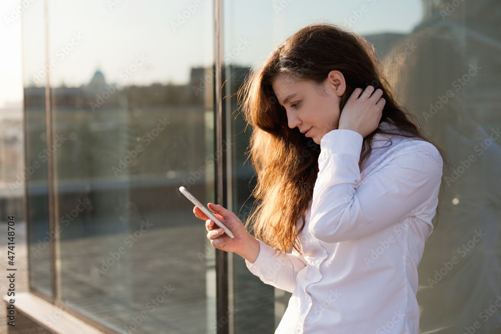 Fototapeta premium young woman texting or video chatting on mobile phone in the city standing near glass office building. Millennial woman using social media on mobile