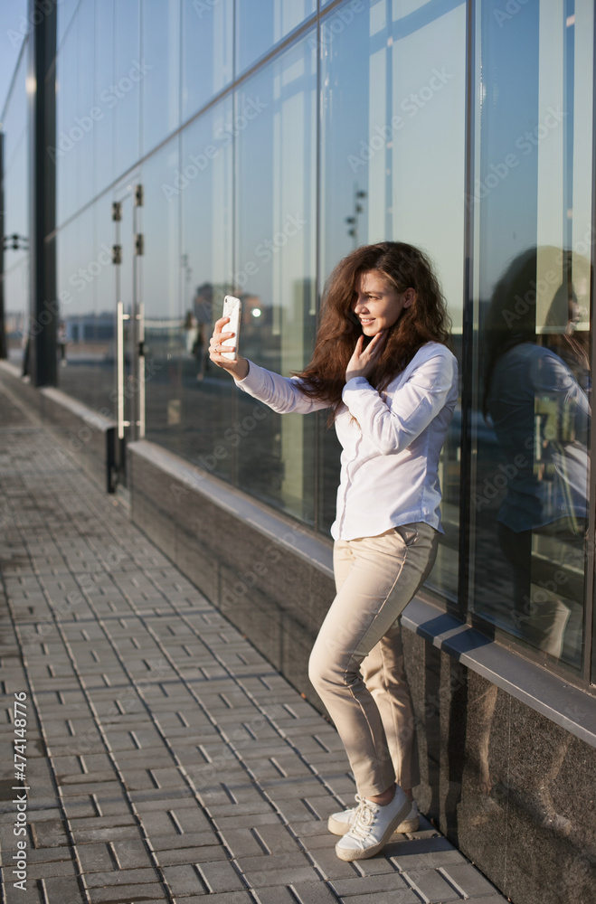 young woman talking or video chatting on mobile phone in the city standing near glass office building