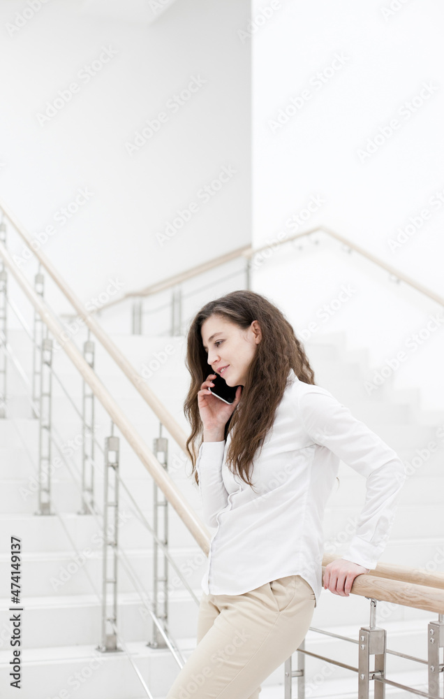 Woman in white shirt using mobile phone standing near white wall. Person using technology in the office stairs. Person using social media or talking on the phone Woman in white shirt using mobile phone standing near white wall. Person using technology in the office stairs. Person using social media or talking on the phone