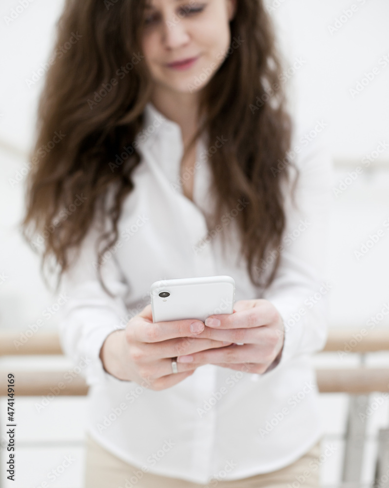 Woman in white shirt using mobile phone standing near white wall. Person using technology in the office stairs. Person using social media or texting Woman in white shirt using mobile phone standing near white wall. Person using technology in the office stairs. Person using social media or texting