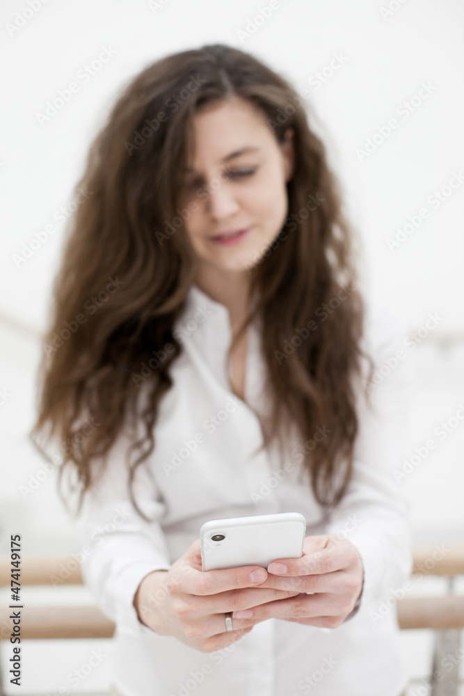 Woman in white shirt using mobile phone standing near white wall. Person using technology in the office stairs. Person using social media or texting Woman in white shirt using mobile phone standing near white wall. Person using technology in the office stairs. Person using social media or texting