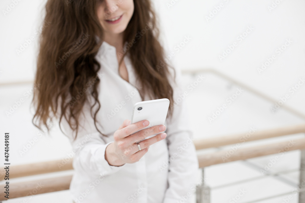 Woman in white shirt using mobile phone standing near white wall. Person using technology in the office stairs. Person using social media or texting Woman in white shirt using mobile phone standing near white wall. Person using technology in the office stairs. Person using social media or texting
