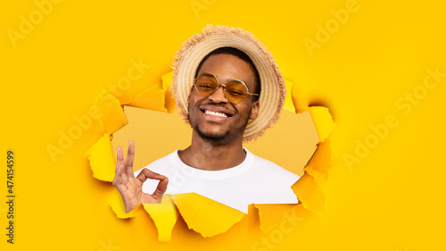 Cheerful relaxed millennial black guy in sunglasses and hat looks through hole in yellow paper and shows ok sign © Prostock-studio