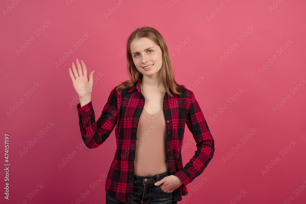 A friendly, young and attractive blonde Caucasian girl in a shirt waving, greeting and looking at camera on a pink studio background.