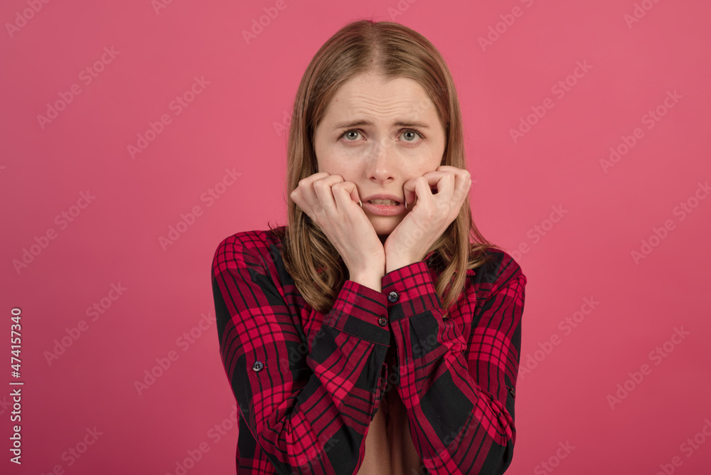 A young and attractive Caucasian blonde girl in a shirt holding her hands on her face, she is scared, isolated on a pink studio background. A young and attractive Caucasian blonde girl in a shirt holding her hands on her face, she is scared, isolated on a pink studio background.