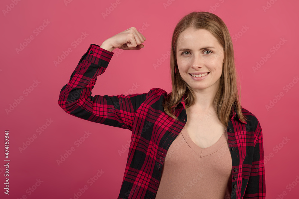 A young and attractive blonde Caucasian girl in a shirt showing her strong hand on a pink studio background. A young and attractive blonde Caucasian girl in a shirt showing her strong hand on a pink studio background.