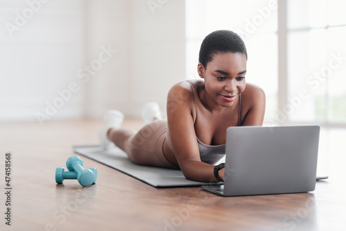 Smiling sporty black woman lying on yoga mat using laptop