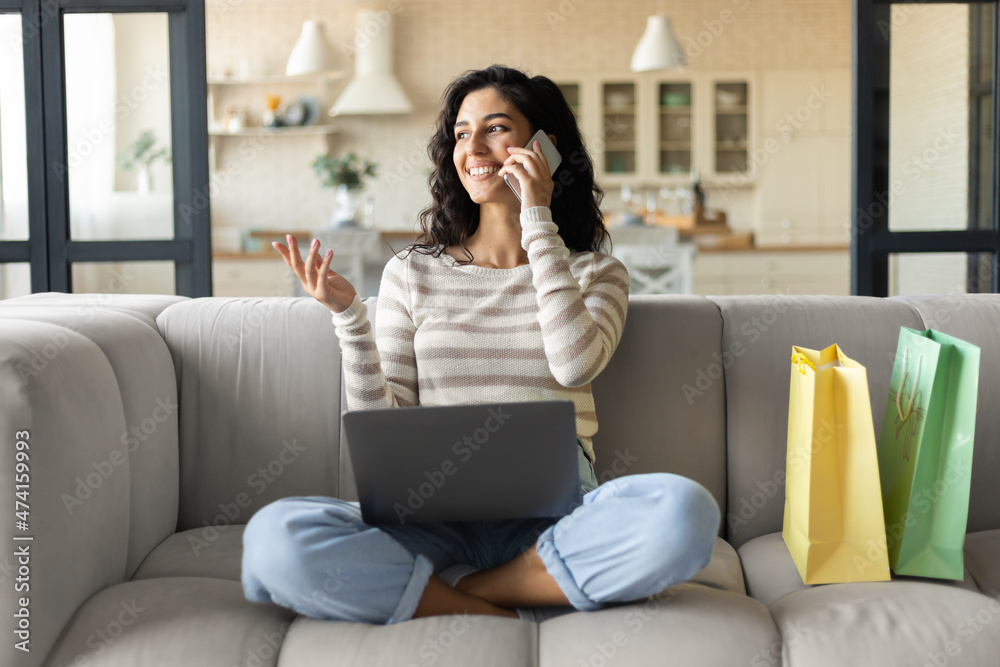 Lovely young brunette woman with gift bags shopping online on laptop, confirming web order on smartphone from home Lovely young brunette woman with gift bags shopping online on laptop, confirming web order on smartphone from home