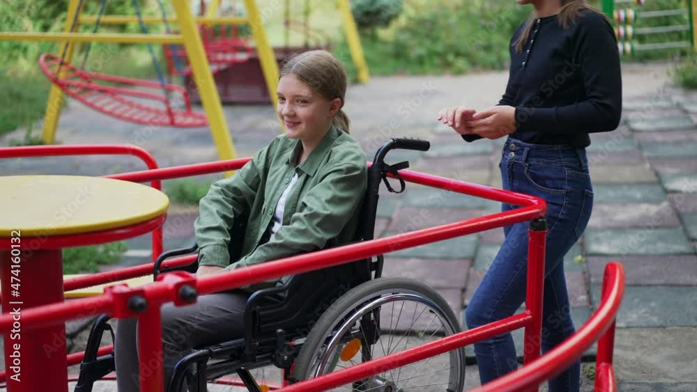 Cute positive Caucasian disabled girl in wheelchair on merry go round with friend standing smiling. Confident supported child resting on playground outdoors. Childhood and injury Cute positive Caucasian disabled girl in wheelchair on merry go round with friend standing smiling. Confident supported child resting on playground outdoors. Childhood and injury
