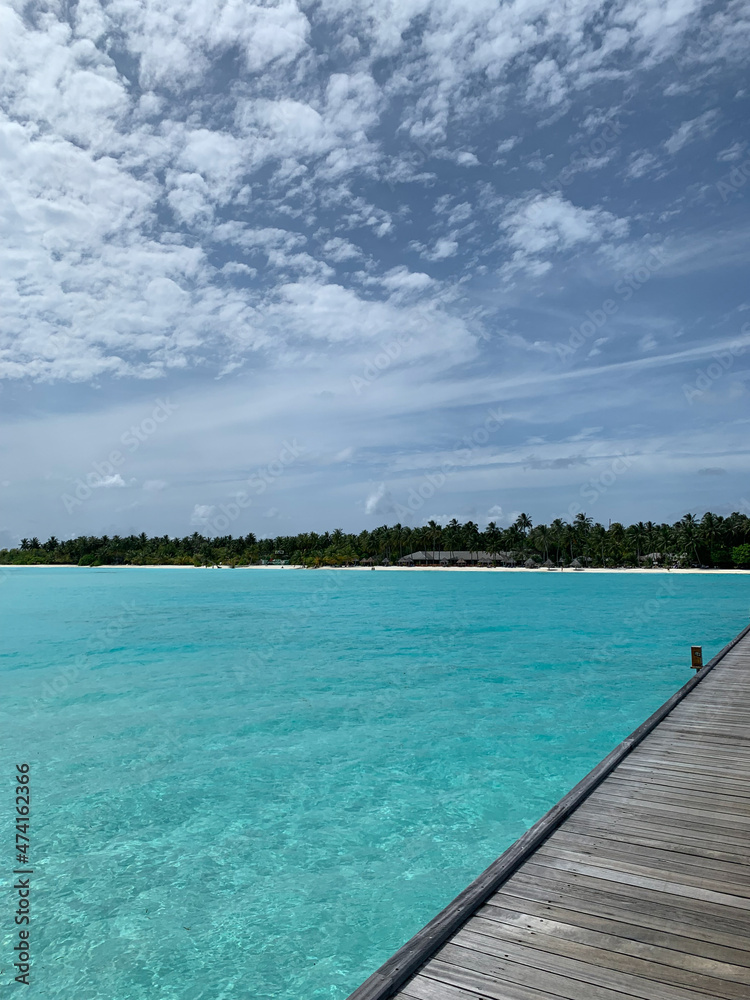 A day in the Maldives. View from a wooden pier to the coast of the Indian Ocean with white sand, azure water, vacationers, sun loungers and palm trees