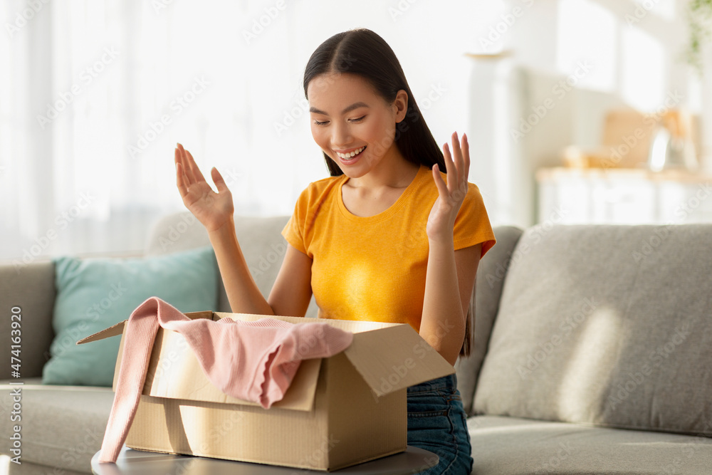 Unpacking clothes. Portrait of happy asian lady opening cardboard box, taking out new shirt, sitting on sofa at home Unpacking clothes. Portrait of happy asian lady opening cardboard box, taking out new shirt, sitting on sofa at home