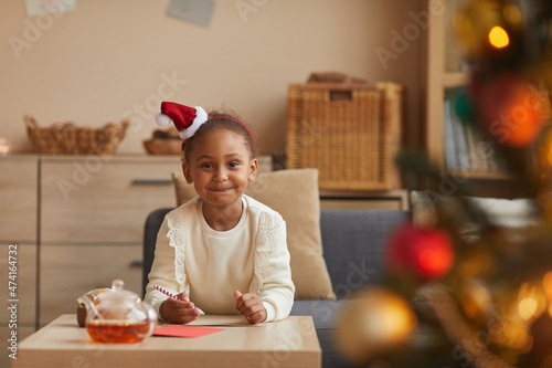 Portrait of cute African-American girl writing letter to Santa while sitting at table by Christmas tree, copy space
