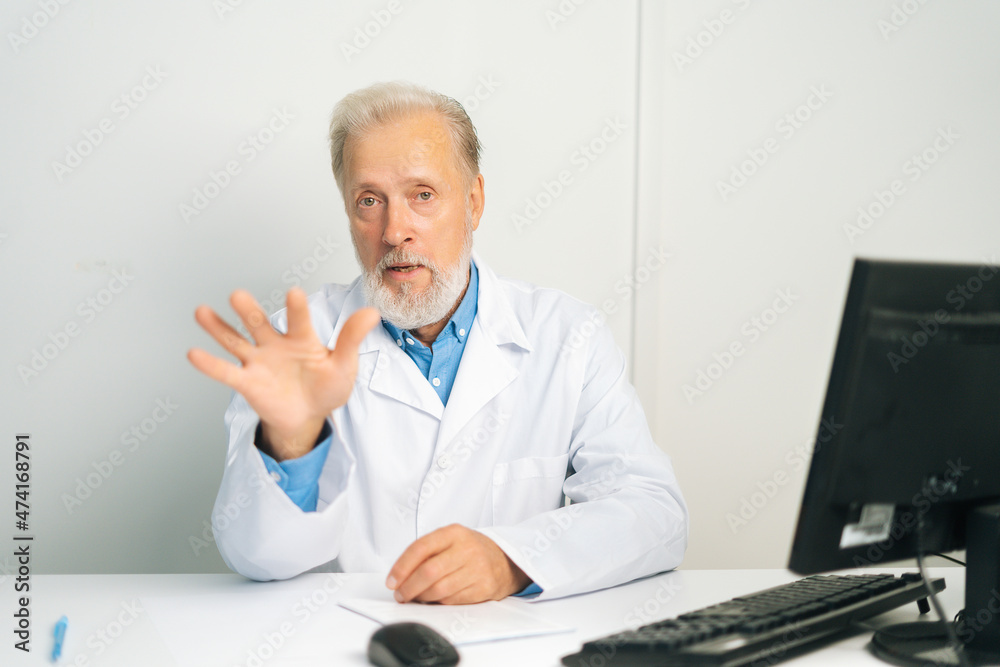 Portrait of professional mature adult male doctor in white uniform coat speaking, looking at camera, gesturing sitting at desk with computer in medical office, consulting patient.