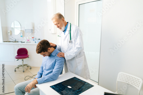 Focused mature adult male manual therapist turning and bending head of male patient to check his injured neck. Young man on preventive examination of doctor chiropractor on neck diagnostic.