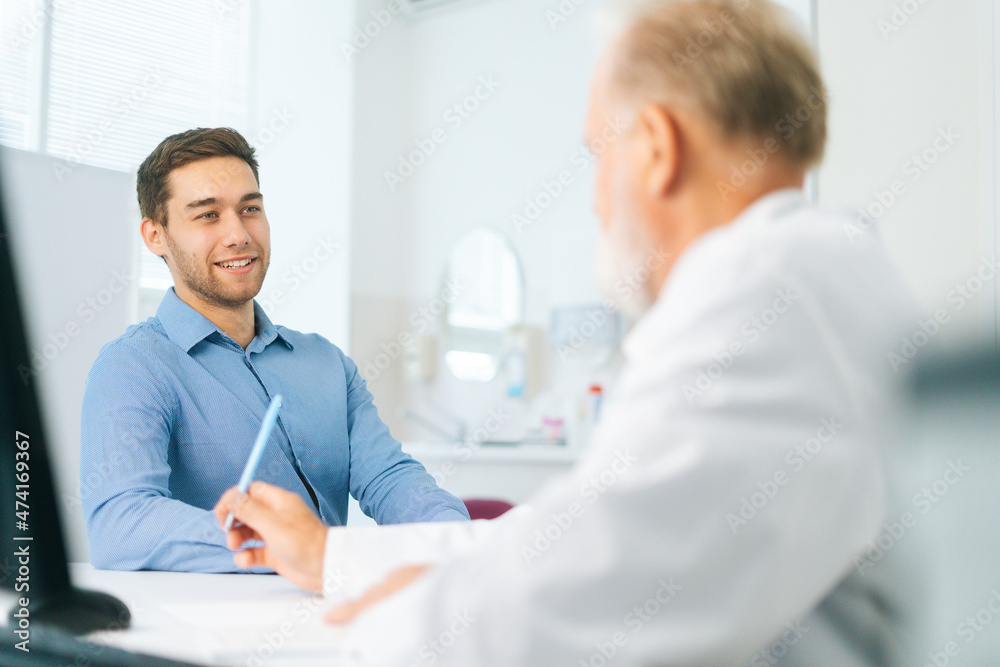 View from back to happy young man patient listening senior male doctor giving healthcare advice during checkup visit in clinic office. Senior physician interviewing smiling young man at hospital. View from back to happy young man patient listening senior male doctor giving healthcare advice during checkup visit in clinic office. Senior physician interviewing smiling young man at hospital.