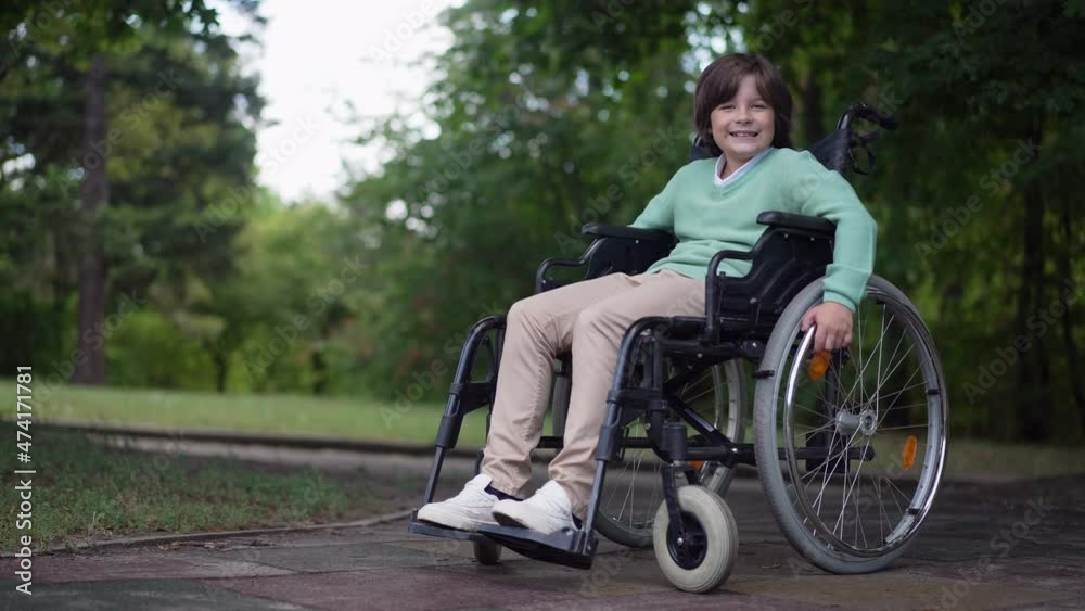 Wide shot happy confident disabled boy in wheelchair looking at camera smiling. Portrait of cute Caucasian paralyzed child posing in summer park outdoors. Confidence and disability Wide shot happy confident disabled boy in wheelchair looking at camera smiling. Portrait of cute Caucasian paralyzed child posing in summer park outdoors. Confidence and disability