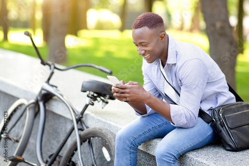 Joyful black man with modern bicycle using cellphone at urban park, copy space