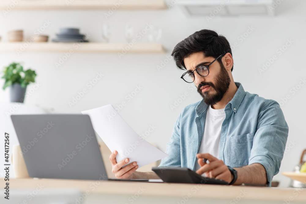 Expensive modern life. Disappointed arab man calculating taxes or planning budget while sitting at kitchen table Expensive modern life. Disappointed arab man calculating taxes or planning budget while sitting at kitchen table