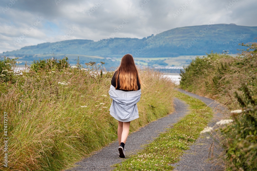 Teenager girl walking on a small country road with amazing view on the ocean and mountain and cloudy sky. County Sligo, Ireland. Knocknarea hill in the background. Travel and tourism concept