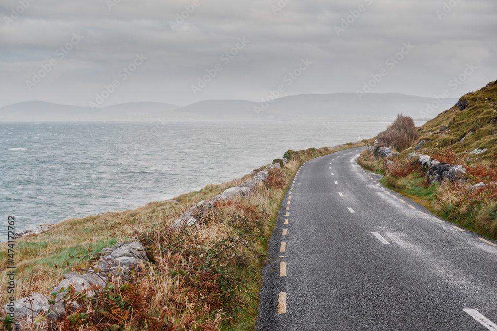 Narrow road by ocean, mountains in the background. Low cloudy sky. Nobody, West of Ireland. Rough terrain. Beautiful nature.