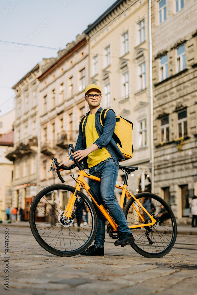 Portrait of a happy delivery man who is sitting on a bicycle and looking at the camera. He is carrying a yellow backpack on his shoulders. Vertical photo.