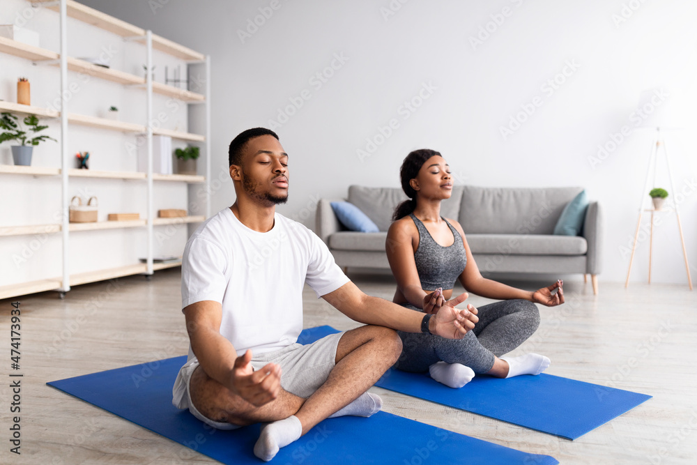 Young black couple meditating with closed eyes, sitting on sports mats in lotus pose at home, full length Young black couple meditating with closed eyes, sitting on sports mats in lotus pose at home, full length