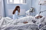 In Hospital, Worrying Young Mistress Sits Beside the Bed where Sick Man Lies. Redhead Woman Is Crying, Hopes for Recovery. In medical Ward Room. Healthcare, People, Human Emotions Concept