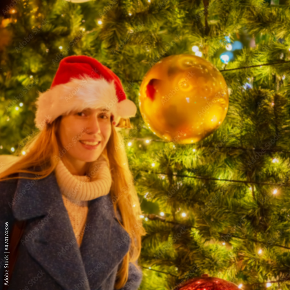 Blurred image of a girl in a Santa hat near a Christmas tree with large balls. Defocused picture