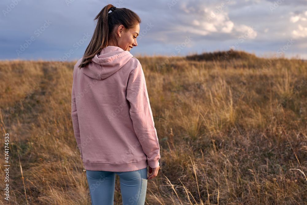 Rear view on happy female in pink hoodie and leggins standing in field in the morning. view from back on long-haired lady going to train on fresh air outdoors, at sunrise. people lifestyle concept