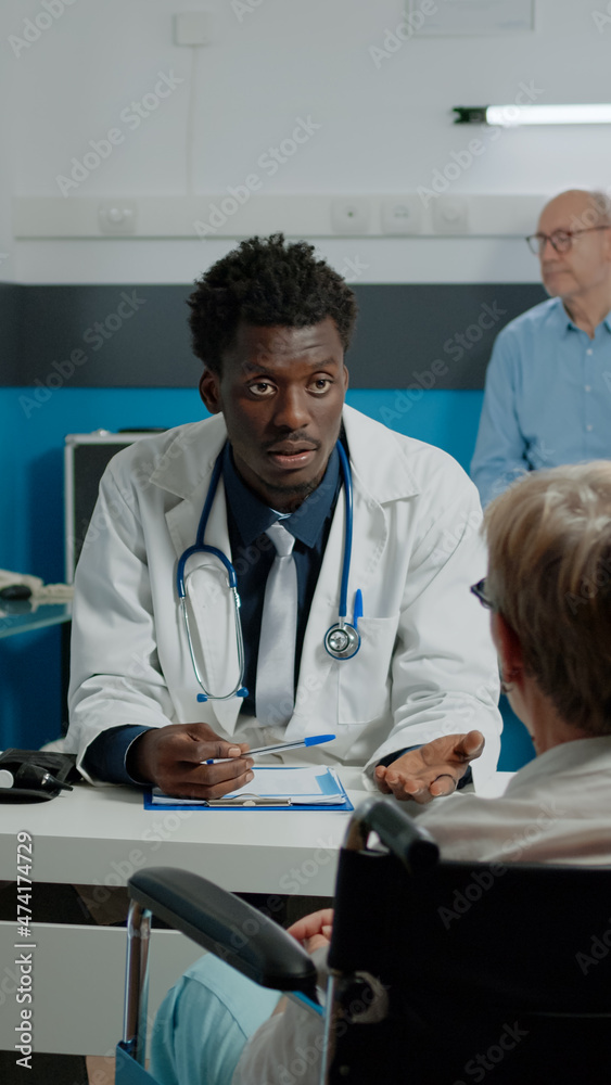 Fototapeta premium Medic discussing about healthcare with disabled old woman at medical facility. Elder patient with handicap sitting in wheelchair while receiving professional consultation from doctor