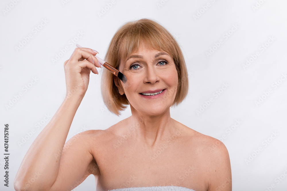 Natural makeup for mature skin. Sexy senior lady putting on decorative cosmetics with brush over light studio background
