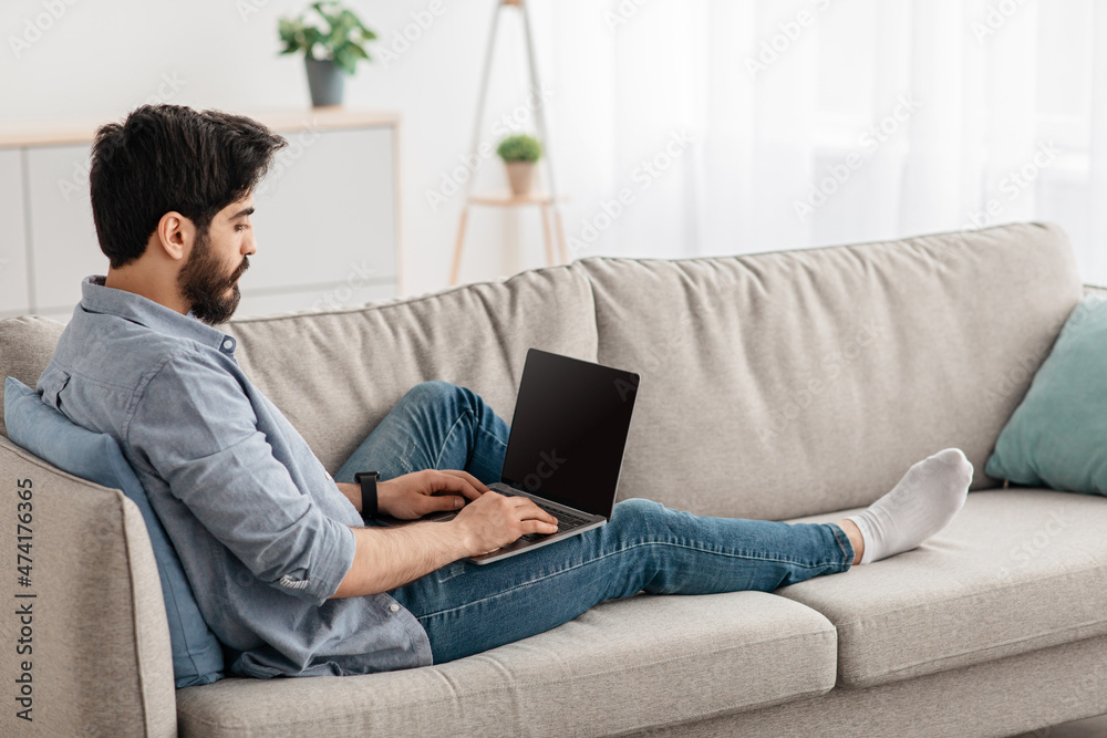Relaxed arab man using laptop with blank screen, resting on couch in living room, home interior, free space, mockup Relaxed arab man using laptop with blank screen, resting on couch in living room, home interior, free space, mockup